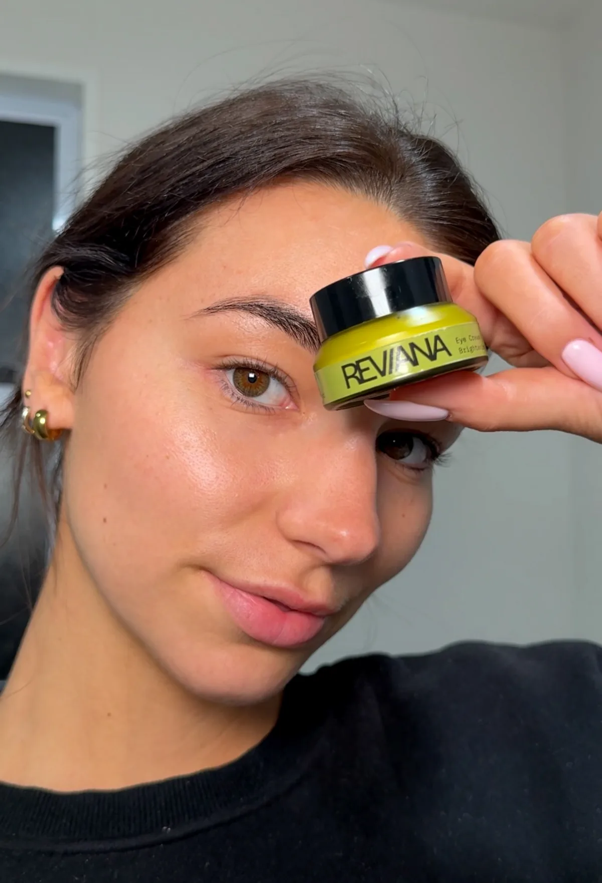 A woman holding the Reviana eye cream jar beside her eye