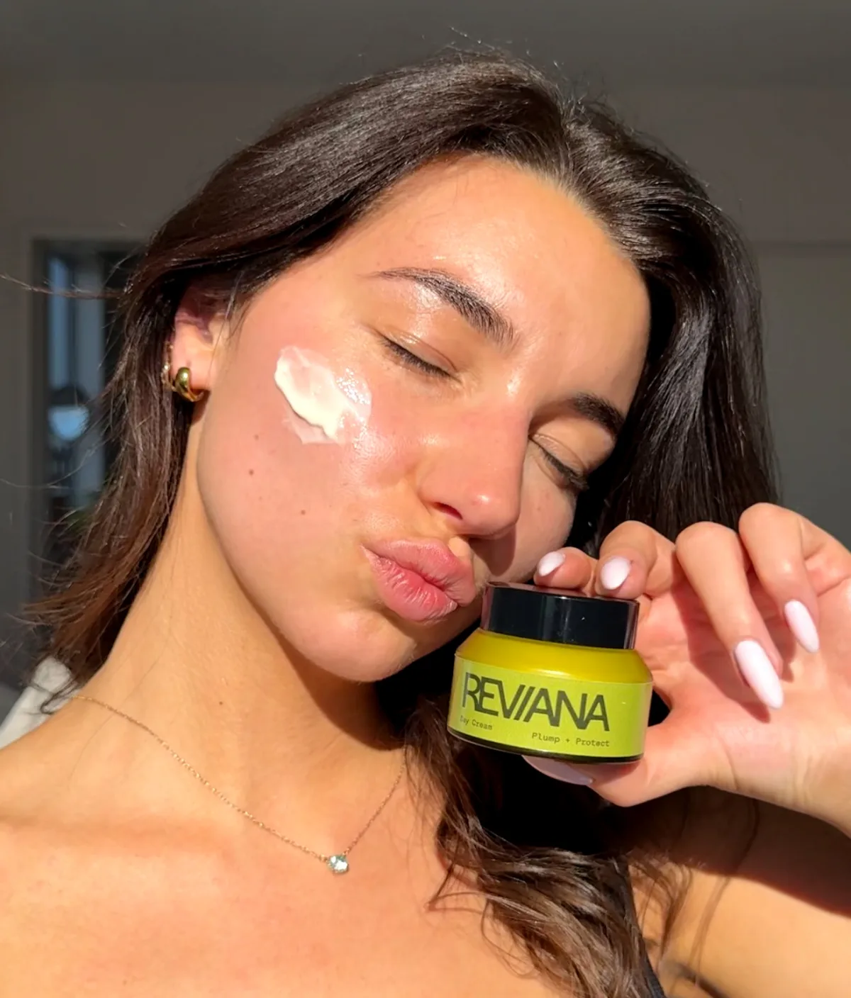 A woman with a swipe of day cream on her cheek, holding the Reviana day cream jar