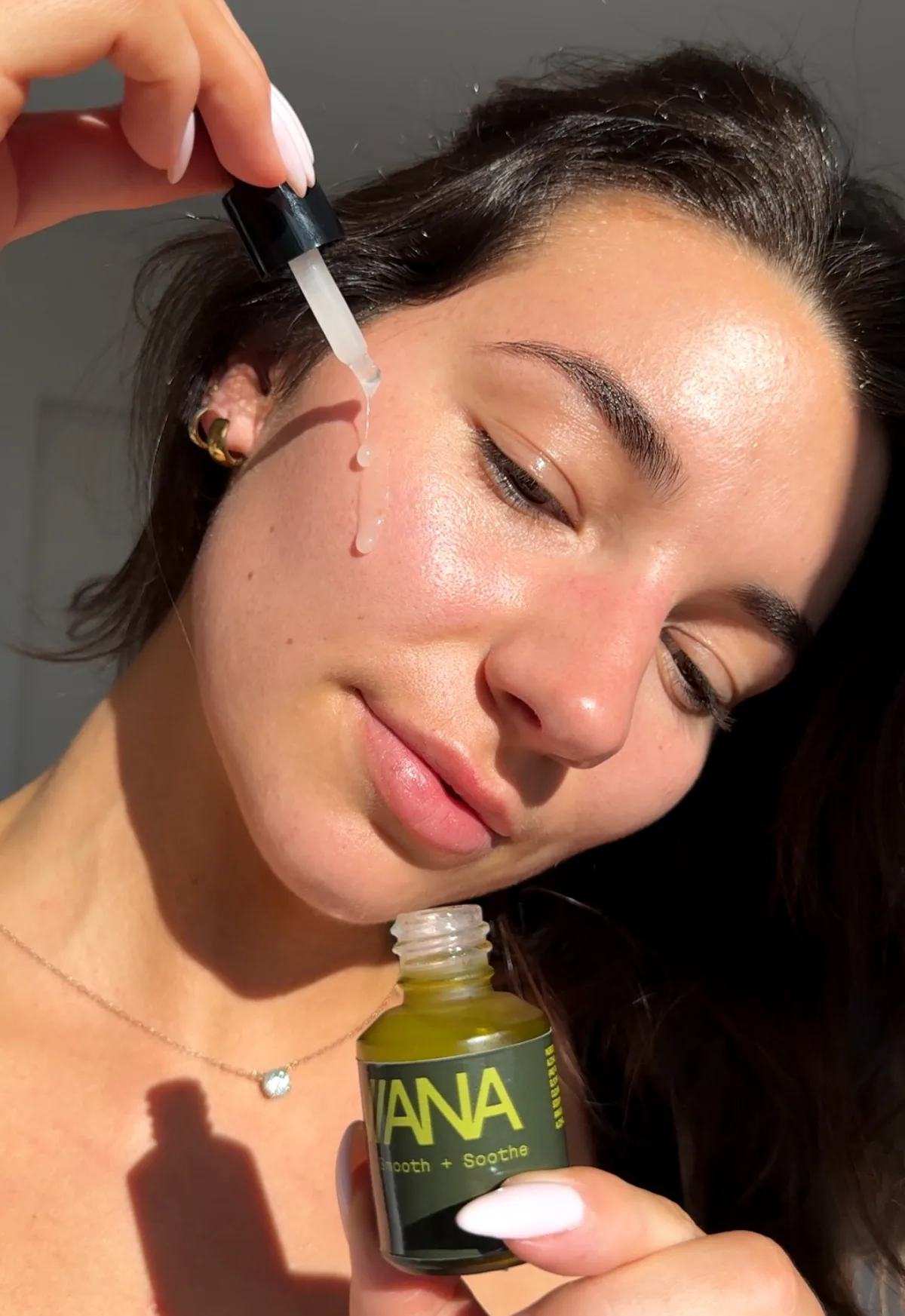 A woman holding the Reviana serum dropper to her cheek in morning light